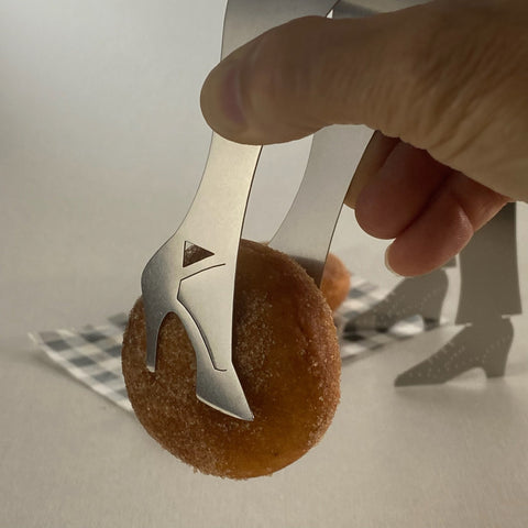 Hand using shoe shaped stainless steel serving tongs to pick up small donut with another set of shoe shaped tongs standing in the background.