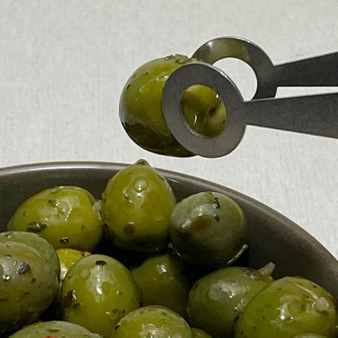 Close up of green olives being picked up by stainless steel serving tongs with circle design on ends