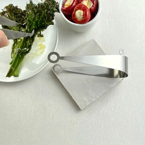 Two stainless steel serving tongs, one lying on white ceramic tile and one picking up brocollini next to bowl of red peppers