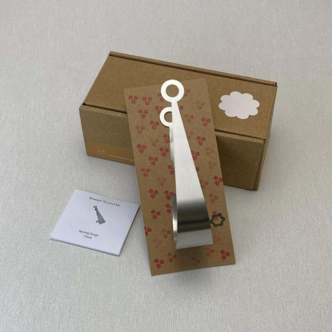 Stainless steel serving tongs in floral stamped cardboard insert leaning on gift box next to illustrated care slip