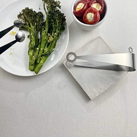 Two stainless steel serving tongs in circle and dome design with plate of brocollini and bowl of mini peppers