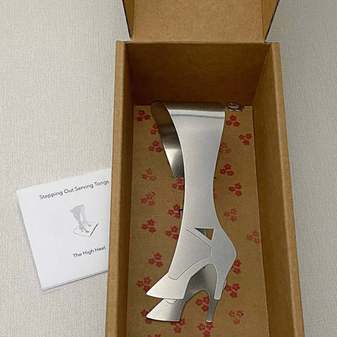 Stainless steel serving tongs in high heel design lying in cardboard gift box with illustrated care slip