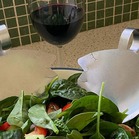 Close up of Those Two stainless steel salad servers with green salad and glass of wine in the background.