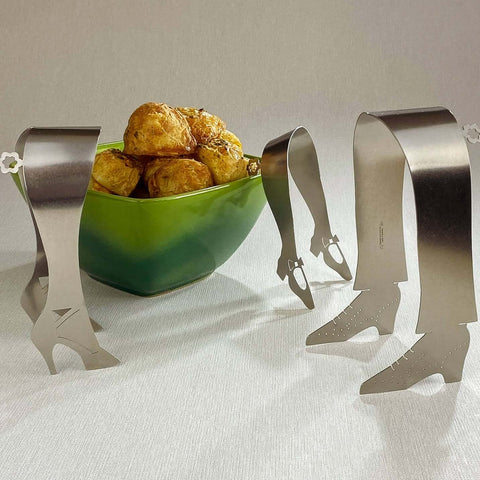 Three stainless steel shoe shaped serving tongs around a green bowl with roast potatoes