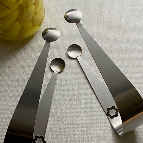 Two pairs domed stainless steel hand crafted serving tongs with jar of yellow pickles in background