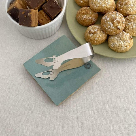 Stainless steel mini tongs lying on blue ceramic coaster next to two plates of biscuits on light grey background