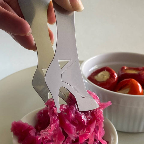 Hand using stainless steel serving tongs in high heel design to pick up pink sauerkraut from bowl with bowl of stuffed peppers in background
