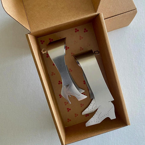 Two stainless steel serving tongs in a shoe shaped design lying in cardboard gift box
