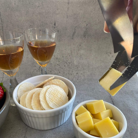 Person using shoe shaped Stepping Out stainless steel tongs to pick up cube of cheese with crackers and glasses of wine in the background