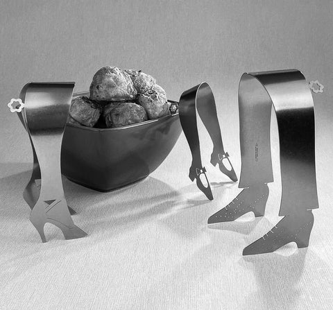 Three stainless steel serving tongs shaped like shoes standing next to bowl of roast potatoes in black and white