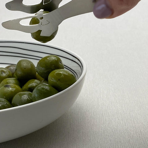 Stainless steel mini tongs shaped like shoe playfully picking up olive with heel from bowl of olives