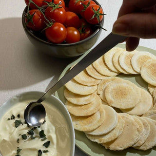 Hand using handcrafted stainless steel tiny serving ladle with yoghurt, rice crackers and cherry tomatoes, with text reading “Time spent is luxury. There’s a pleasure in hand crafted that can’t be replicated”