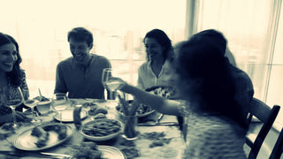 Friends sharing a relaxed meal around a table with wine, food and handcrafted serving pieces with text that reads "Pieces that belong together. Servingware sets. Beautifully boxed and ready to give".