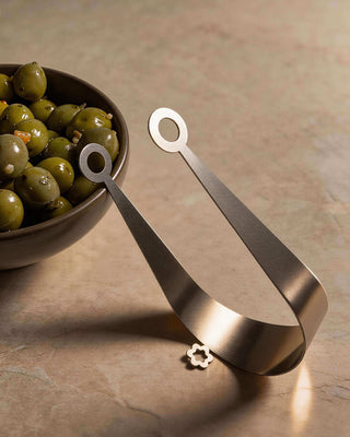 Circles handcrafted stainless steel serving tongs beside bowl of marinated green olives on stone surface