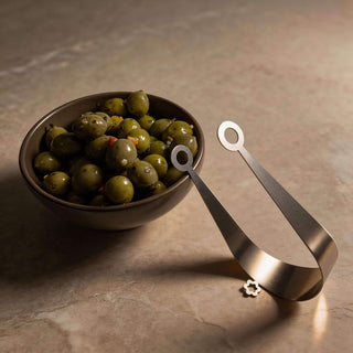 Handcrafted Circles stainless steel serving tongs beside bowl of marinated green olives on stone surface
