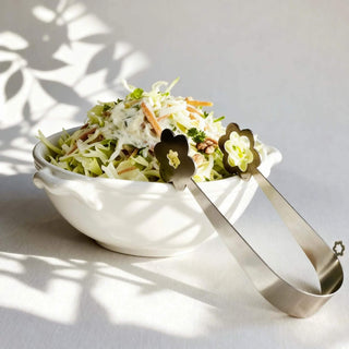 Handcrafted stainless steel Kaleidoscope serving tongs beside bowl of coleslaw salad in soft natural light