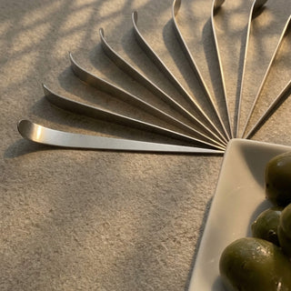 Stainless steel Maestro cocktail picks arranged in a fan shape beside green olives on a white dish