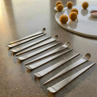 Set of handcrafted stainless steel Maestro cocktail picks beside arancini balls on a white serving platter