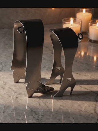 Video of the Stepping Out handcrafted stainless steel serving pair standing on a marble surface in soft candlelight, showing the sculptural high heel design and smooth metal finish.