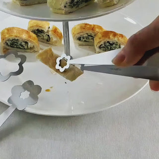 Video of the Flowers handcrafted stainless steel serving pair lifting a small savoury pastry from a white serving plate, showing the flower-shaped detail.