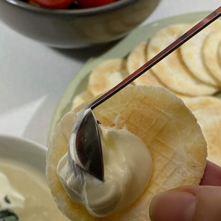 Stainless steel tiny serving ladle spreading yoghurt onto a crisp cracker beside cherry tomatoes